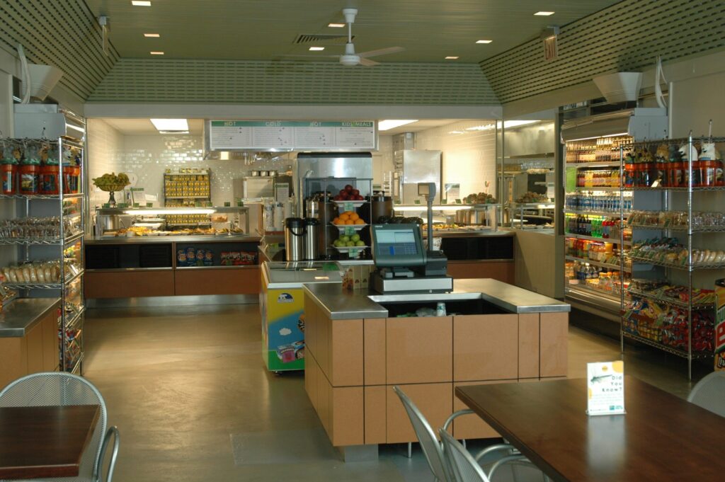 Modern cafeteria with food display counter and a chef at work in the kitchen area.