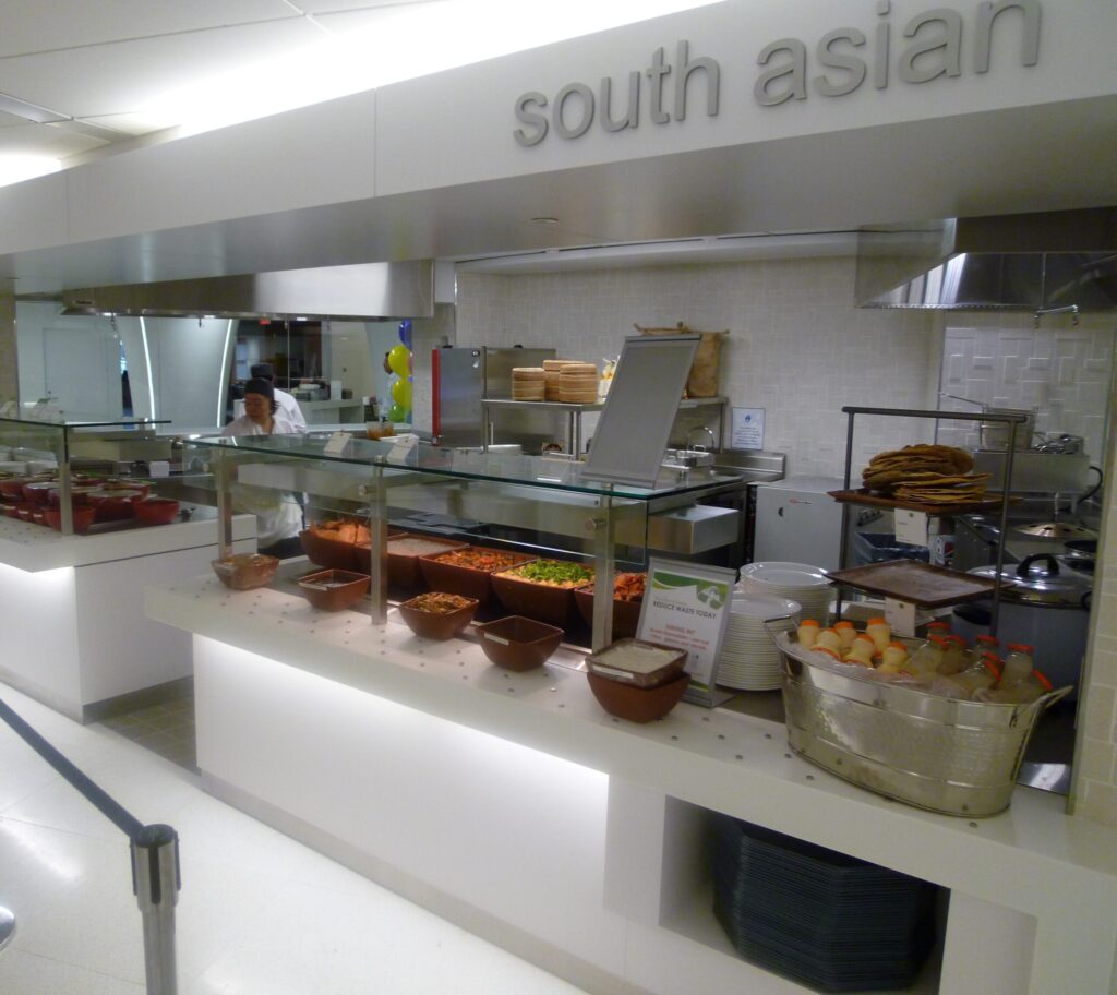 Modern cafeteria with food display counter and a chef at work in the kitchen area.