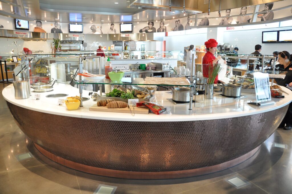 Modern cafeteria with food display counter and a chef at work in the kitchen area.