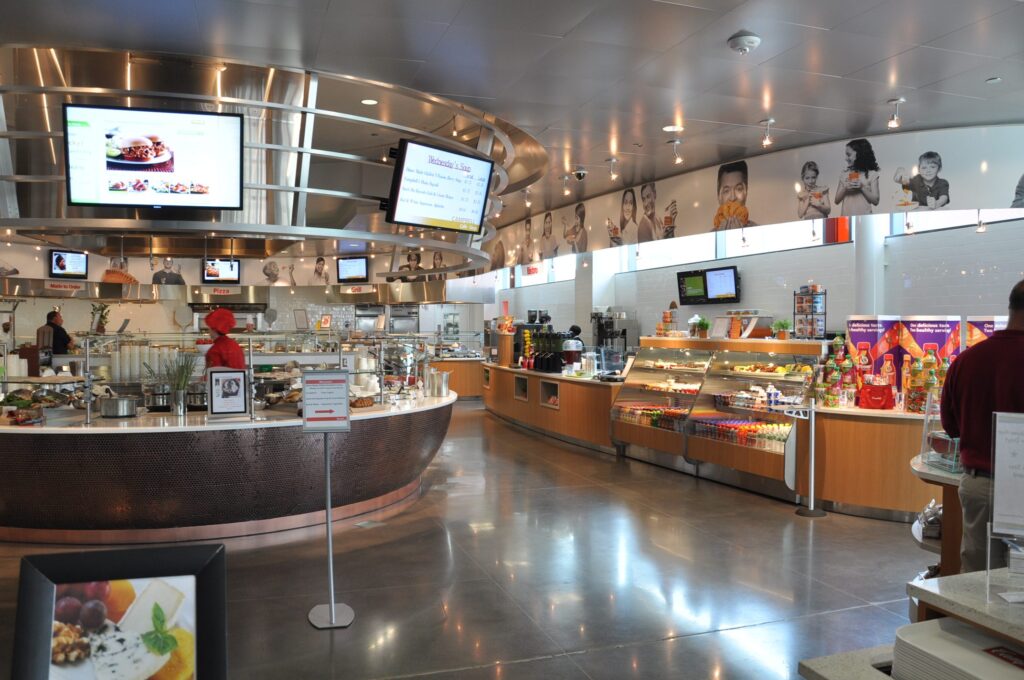 Modern cafeteria with food display counter and a chef at work in the kitchen area.