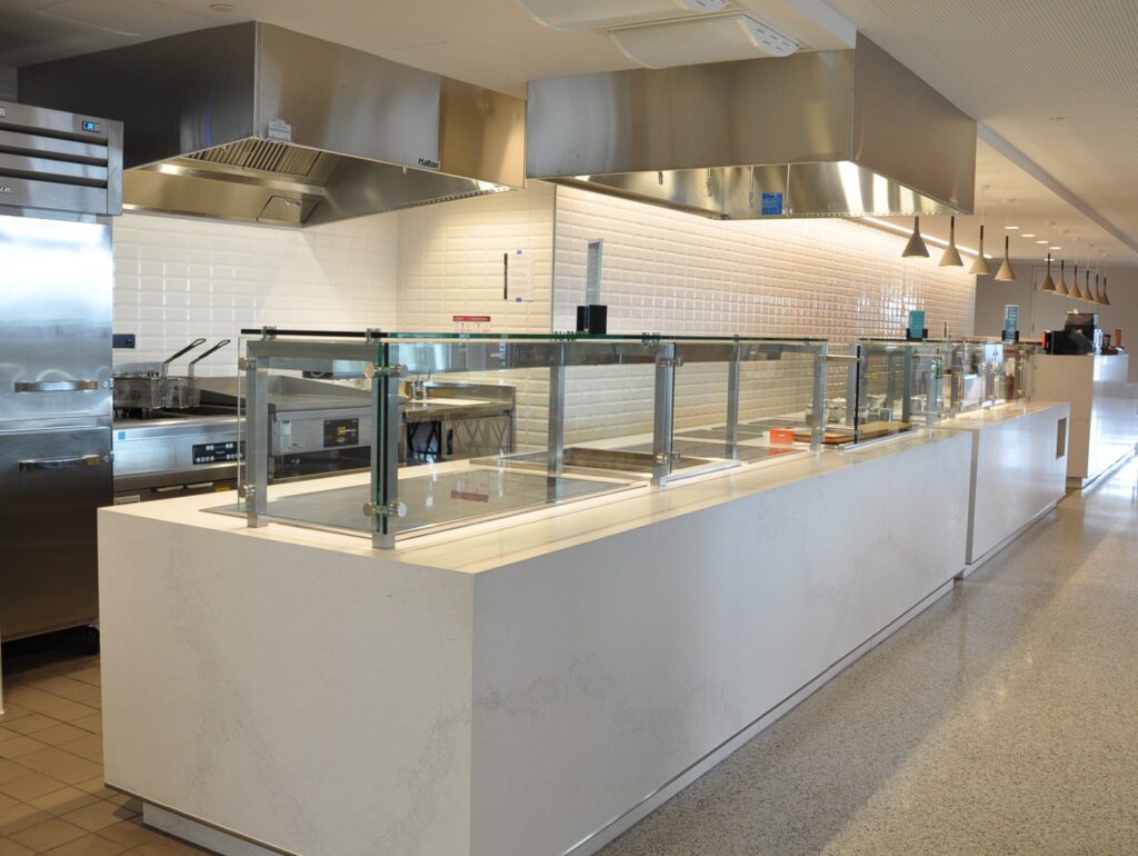 Modern cafeteria with food display counter and a chef at work in the kitchen area.