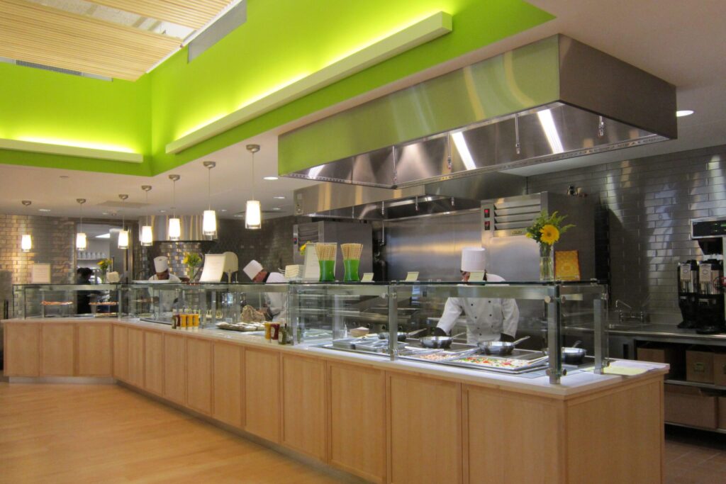 Modern cafeteria with food display counter and a chef at work in the kitchen area.