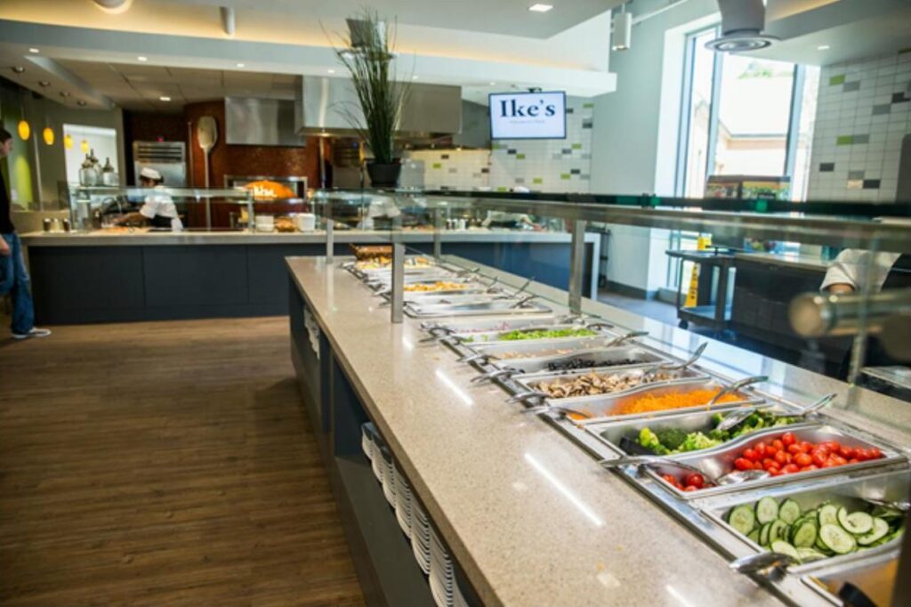 Modern cafeteria with food display counter and a chef at work in the kitchen area.