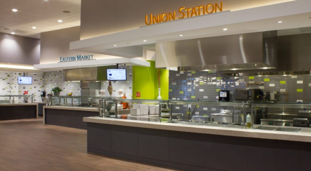 Modern cafeteria with food display counter and a chef at work in the kitchen area.